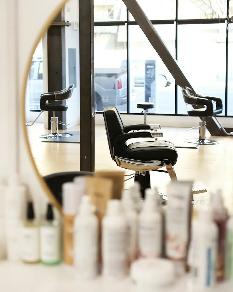 A black styling chair beside a round mirror, with bottles on a shelf in the foreground