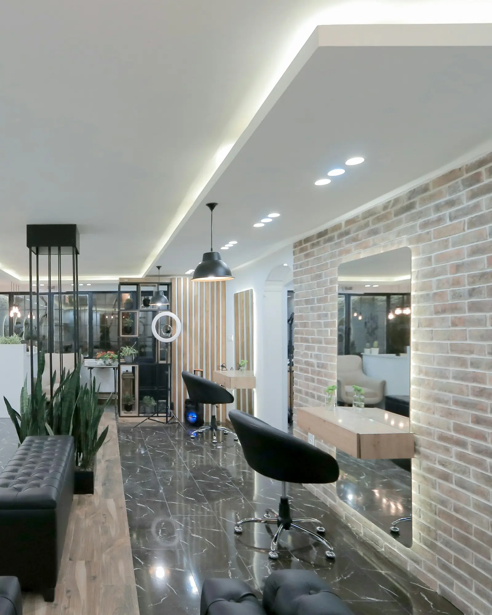 Inside the salon: black styling chairs facing round lit mirrors, with a brick wall and plants