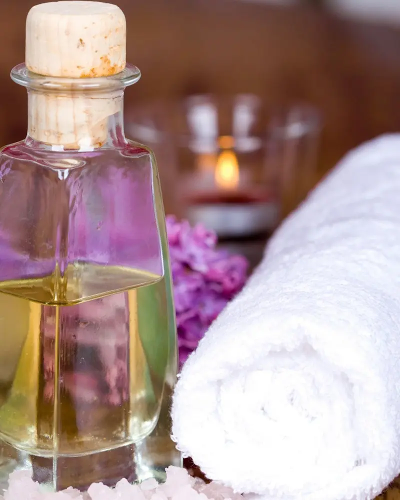 A glass bottle of massage oil beside a rolled white towel and bath salt