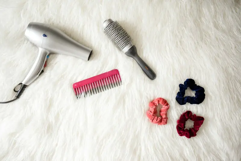 A hair dryer, round brush and pink comb on a soft white rug
